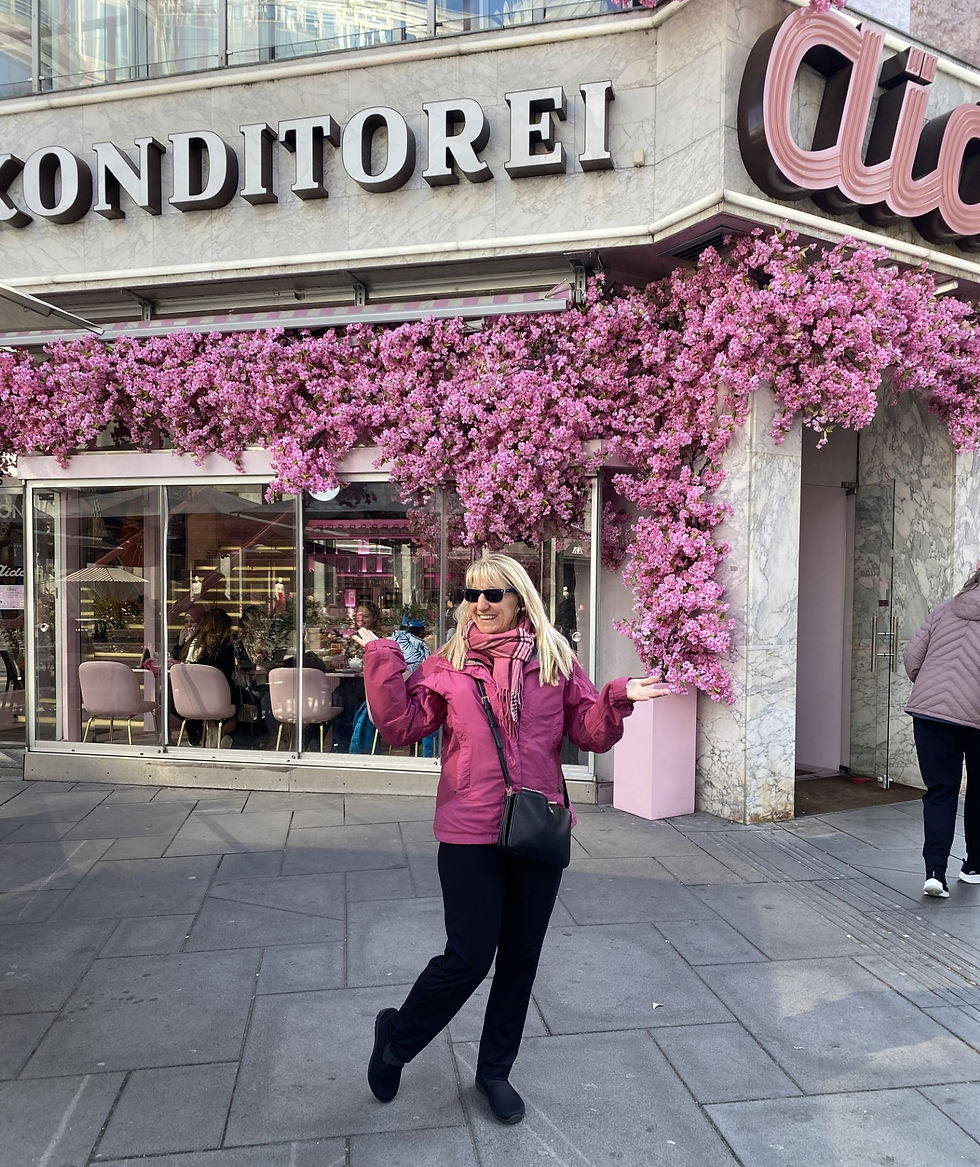 Smiling woman in pink jacket poses outside Conditorei café, framed by pink blossoms and glass windows on a city sidewalk.