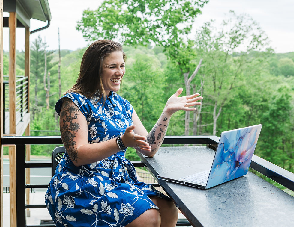 Smiling woman in a blue floral dress gestures beside a laptop on a balcony, with green trees in the background.