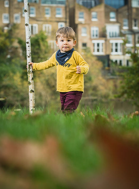 A polished portrait of a child in a yellow sweater and purple trousers, holding a birch tree in front of London heritage buildings, capturing a sense of quiet continuity.