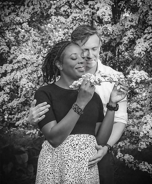 A romantic black and white outdoor maternity portrait by Pennypics - a couple embracing amidst spring blossoms, featuring a mother-to-be gently holding her belly while her partner looks on with sincere care.