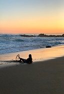 A beach, sunset, sky of oranges, yellows and blues. A silhouette of a young girl lying on the shore where the waves crash, kicking up the water.