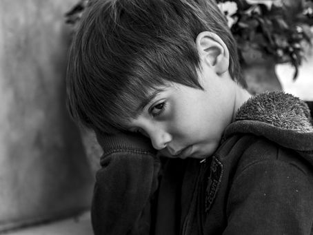 A sad child in a hoodie leans against a wall, resting their head on their hand. The background is blurred, featuring leaves.