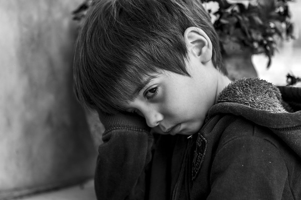 A sad child in a hoodie leans against a wall, resting their head on their hand. The background is blurred, featuring leaves or flowers.