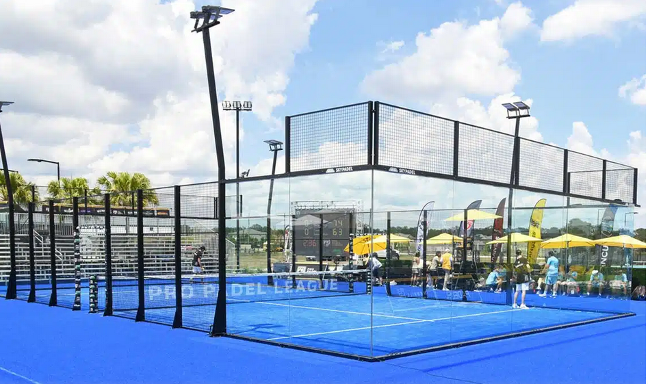 Outdoor super panoramic padel court with a fully glazed enclosure and no visible corner posts, featuring vibrant blue turf and sleek black framing. Located in a professional tournament setting with grandstands, flags, and lighting poles, this court offers maximum visibility for spectators under a partly cloudy sky.
