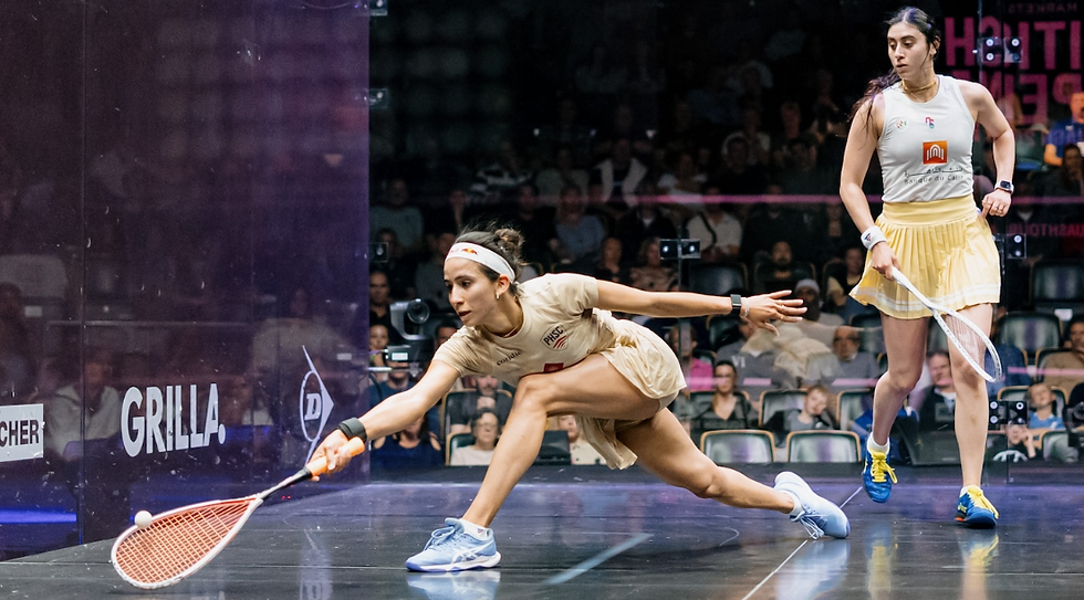 Two professional female squash players during a match. One player lunges low to hit the ball near the front wall while the other watches from behind. The court is glass-walled, and spectators are visible in the background.