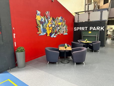 Lounge area with modern chairs and tables in front of a red wall with a colorful sports-themed mural, located inside the Sportpark.