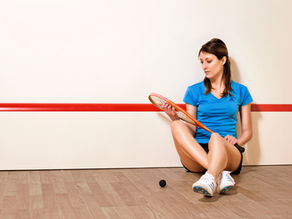 Woman in blue shirt, sitting on a squash court floor, holding a racket.