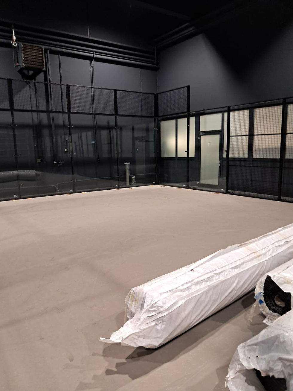 Indoor padel court under construction with a bare floor, black metal fencing, glass walls, and rolls of artificial turf wrapped in plastic on the ground.