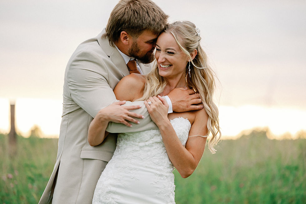 Bride in white gown smiles as groom in tan suit hugs and kisses her in a grassy field at sunset, warm and joyful.