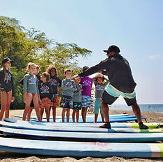 GotʻUm Surfing School Owner Lopaka Samudio teaching surf lessons stands in position on his feet showing how to stand up on a surfboard to kids on a beach in Lahaina Maui