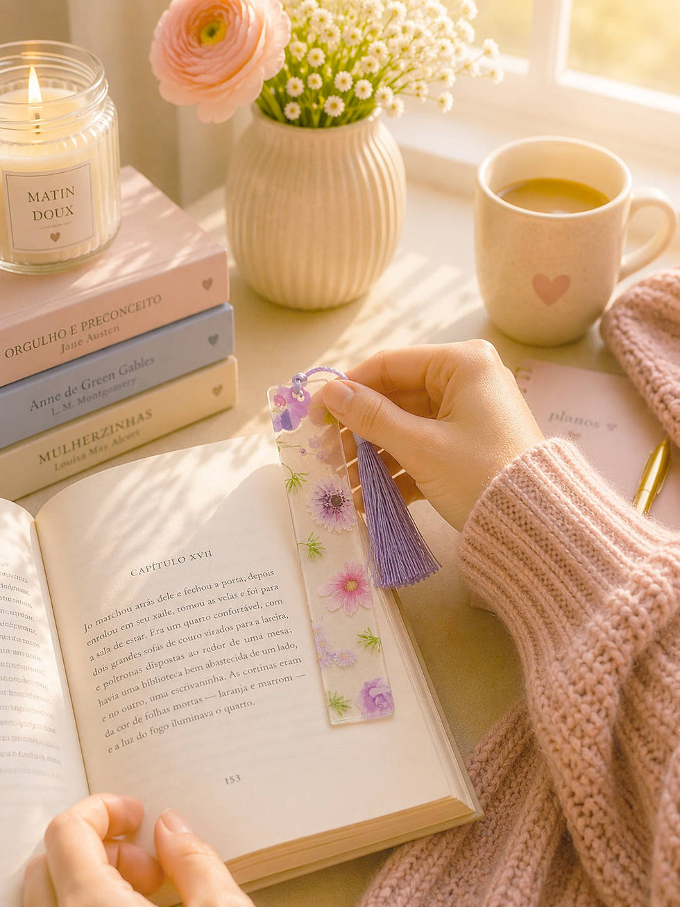 Mão marcando a página de um livro aberto com marca-páginas de resina floral roxo; vela, flores e caneca de café com leite sob luz suave, clima aconchegante.
