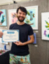 Winner of Most Engaging Artist Award at the 2023 Minnehaha Falls Art Fair, standing in artist booth holding award certificate