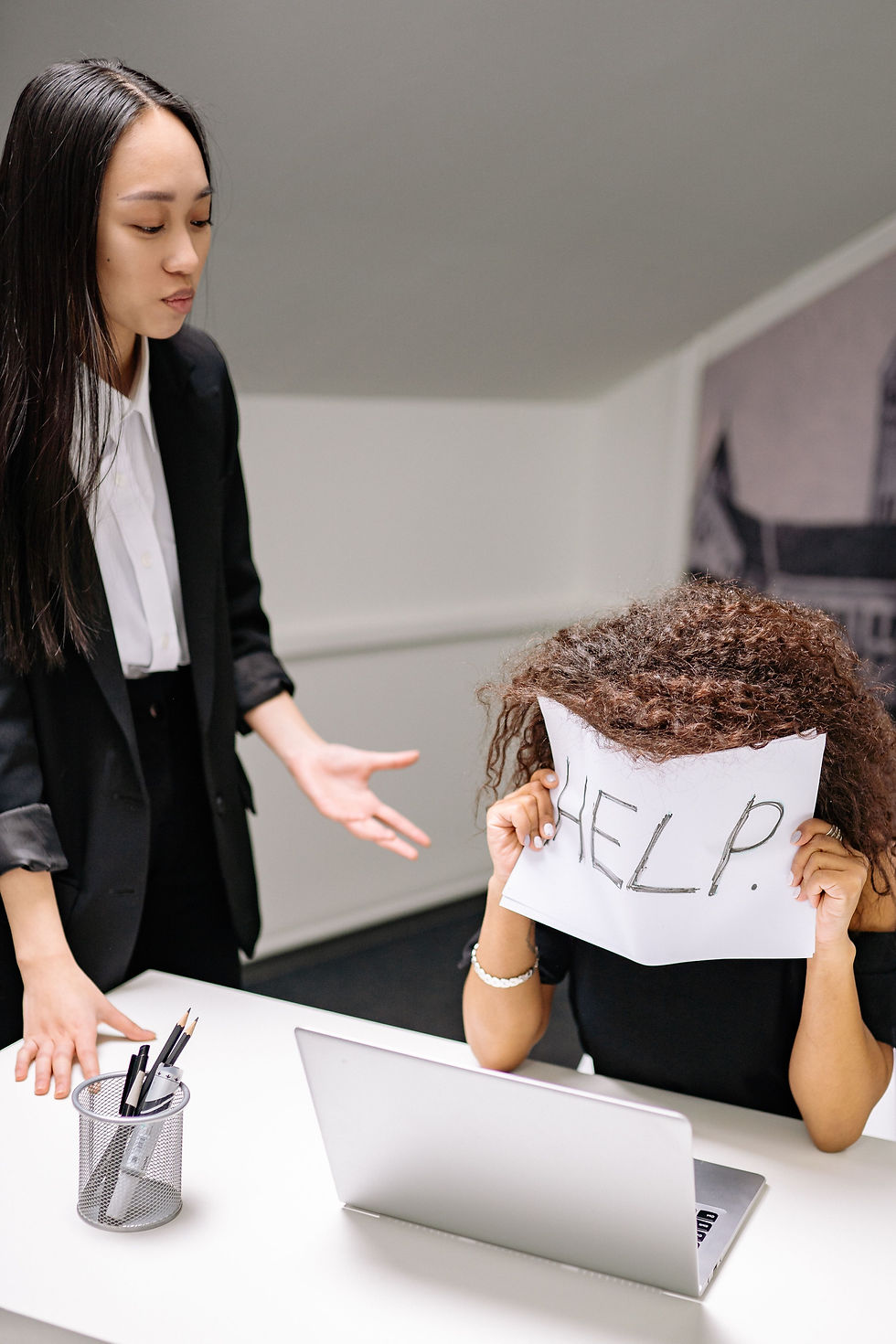 Frau sitzt am Schreibtisch und hält sich Schild mit "Help" vors Gesicht, während andere Frau eine Antwort von ihr erwartet