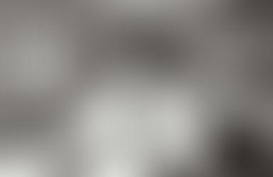 Black and white photograph of Dr. Helen Rodriguez-Trias, a Latina physician and public health advocate, sitting at a desk in a medical office. She is wearing a white lab coat over a turtleneck sweater, smiling warmly while looking at the camera. Papers, books, and a pair of glasses rest on the desk, with shelves of documents in the background.