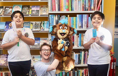 Two young boys stand either side of a man performing a brown and blue puppet character. The children are holding handmade bookmarks proudly. They are in a library setting as part of a workshop.