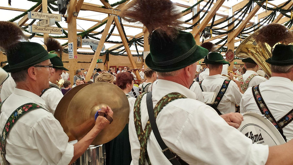 Musicians in traditional Bavarian attire play brass instruments and cymbals in a festive hall with wooden beams and greenery.