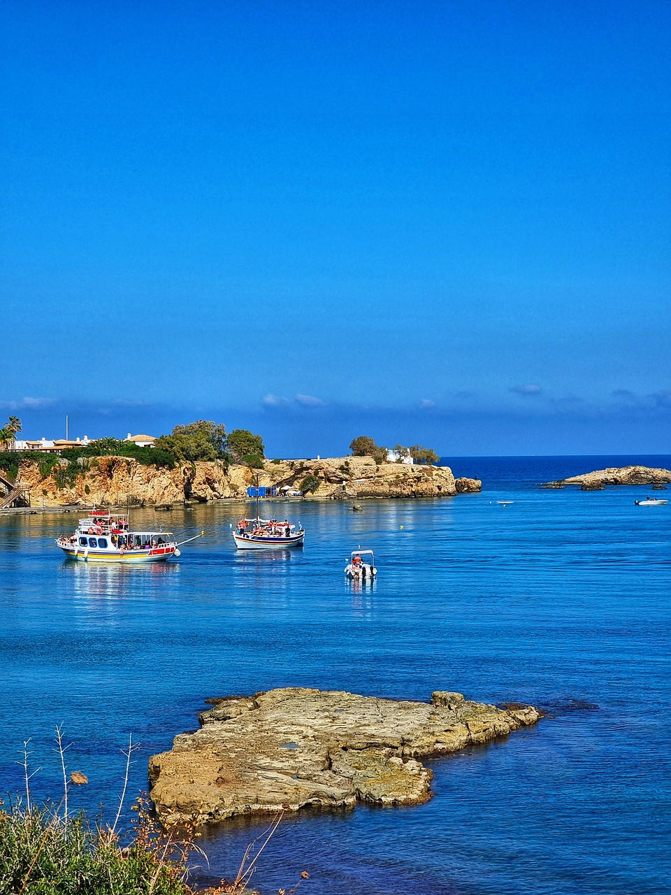 Boats float on a calm, blue sea near rocky shores under a clear sky. Vegetation and cliffs are visible in the background. Relaxing scene.