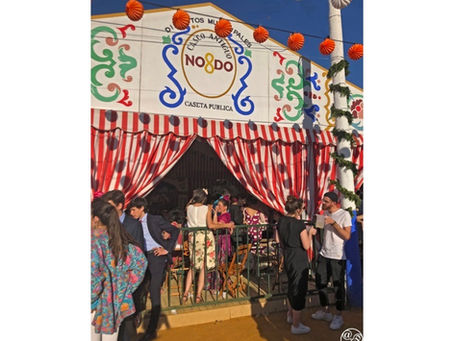 People socializing outside a festive tent with red-striped curtains. Colorful patterns and "NO8DO" text on the facade. Bright, lively atmosphere.