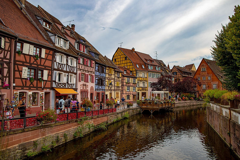 Colorful half-timbered houses line a canal with people walking along the path. Blue sky, vibrant flowers, and a relaxed, charming atmosphere.