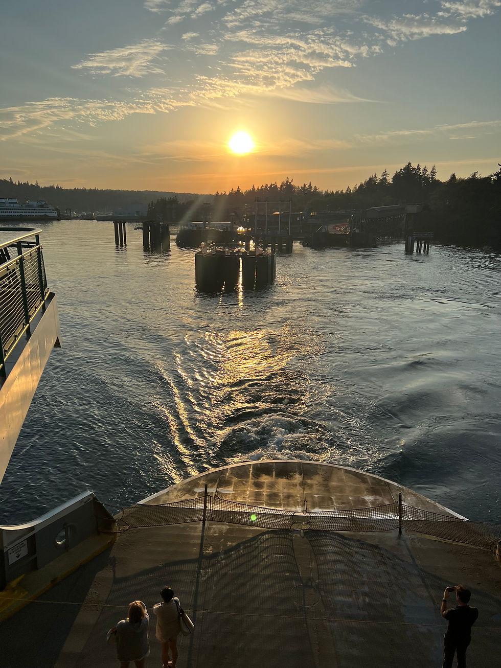 Sunset view from a ferry; three people enjoy the scenery with rippling water and distant trees. Warm tones create a serene mood.