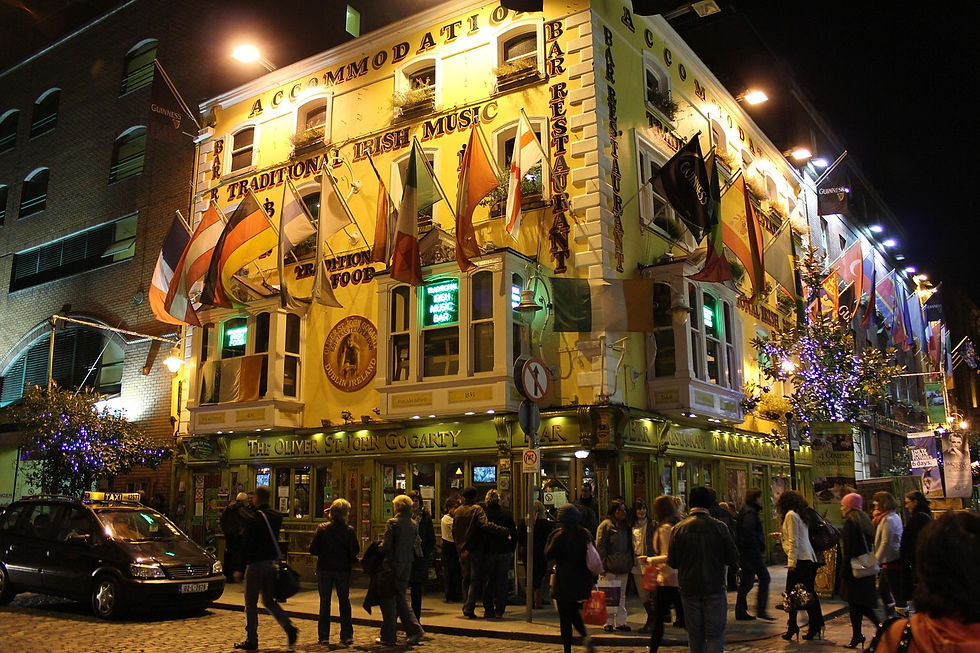 Crowded Dublin street at night with people passing by a brightly lit pub adorned with flags. The building reads "The Oliver St. John Gogarty."