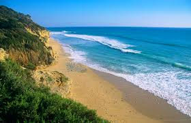 A serene beach scene with clear blue skies, golden sand, gentle waves, and green cliffs. The mood is peaceful and inviting.