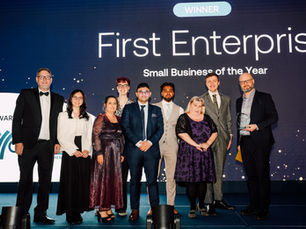 A group of nine businesspeople in formal wear stand on a stage in front of a screen with the text: First Enterprise, Small Business of the Year. A middle-aged man on the right side of the stage is holding a glass award’s trophy.