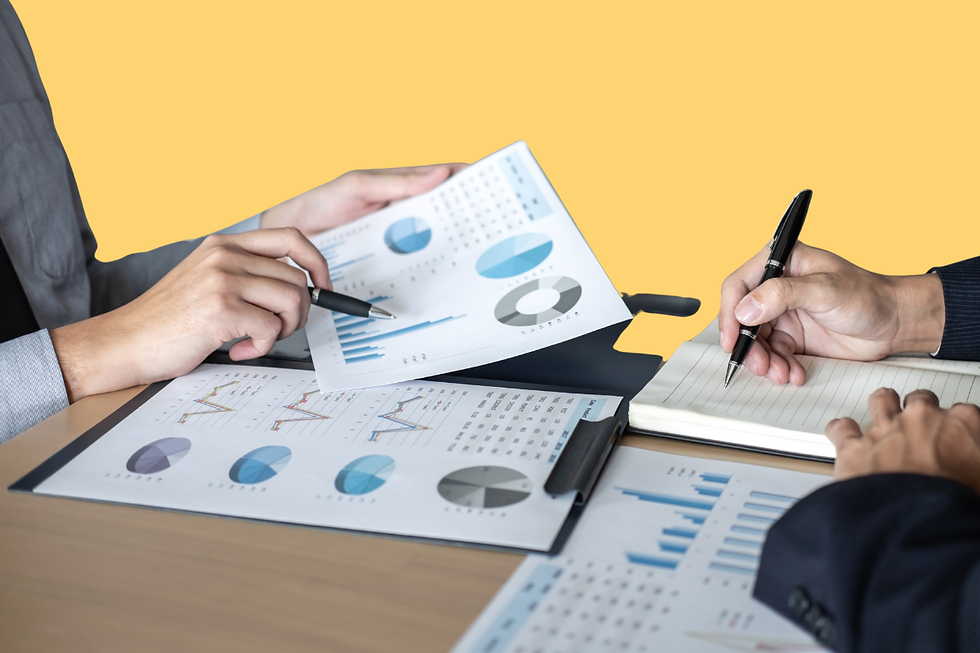 Hands review business charts and graphs while one person takes notes in a meeting, with pens on a table and a yellow background.