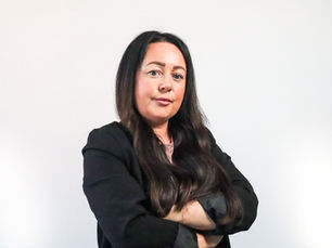 A middle-aged woman with long dark hair (Kimberley Stanley) stands against a dark blue wall facing the camera. She is wearing a black top.