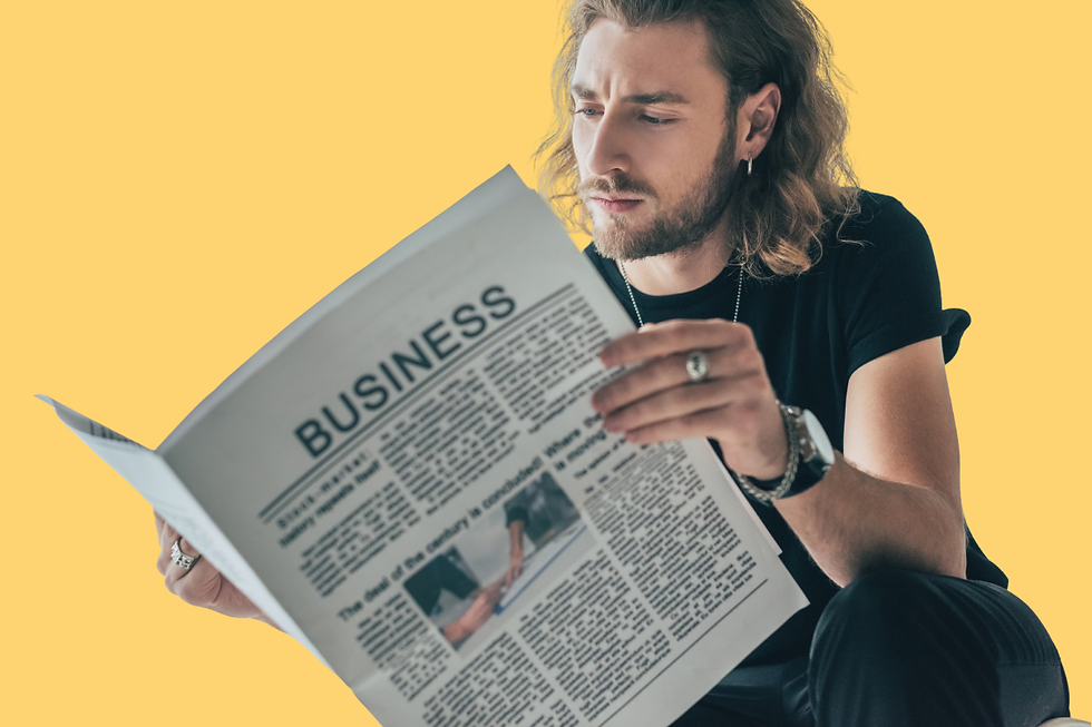 A young man with long hair reads a newspaper with a headline that reads: “Business”, while kneeling in front of a yellow background