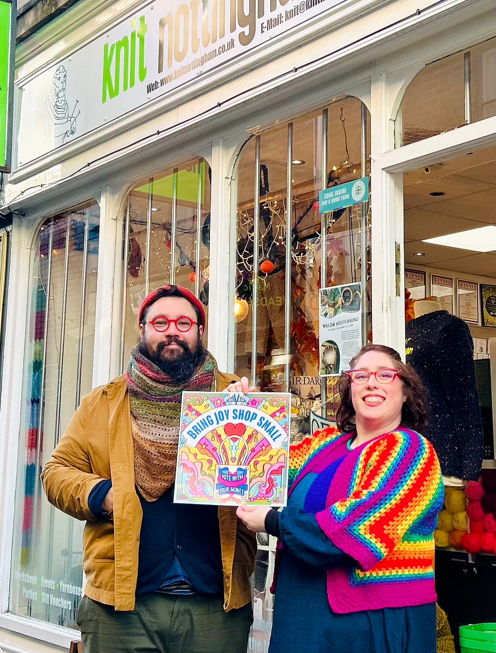 Two people stand outside Knit Nottingham holding a colorful Bring Joy Shop Small sign, wearing bright knitwear and red glasses.