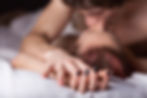 Close-up of a couple kissing in bed, with a hand of black-painted nails in focus against white sheets.