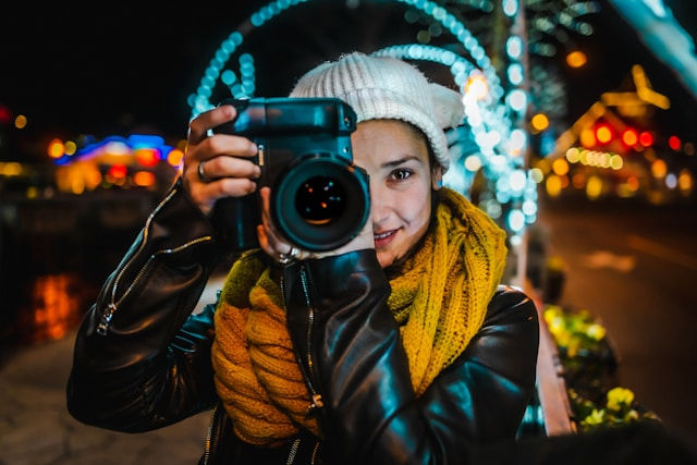 Person in a white beanie and yellow scarf holds a camera, smiling at night. Background features colorful, blurred lights and arches.