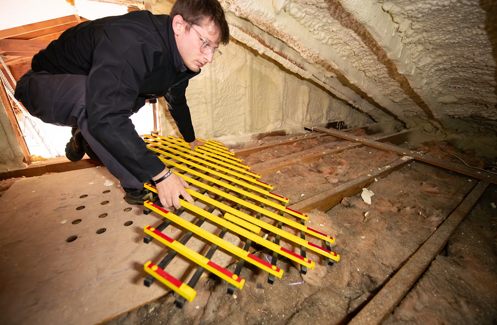 Man in glasses crouches in an attic, adjusting a yellow slatted grate beside spray foam-insulated rafters.