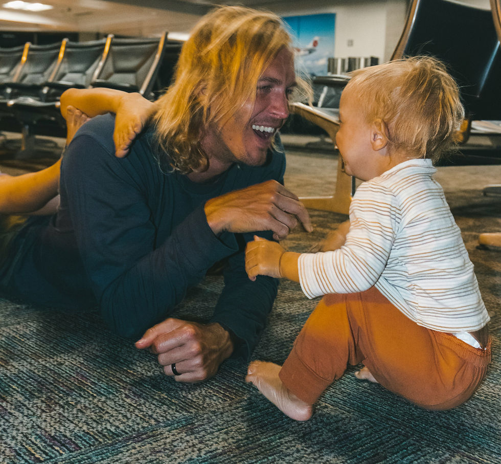 A man with long hair plays and smiles with a toddler on the floor of an airport terminal. The mood is joyful with overhead lights.