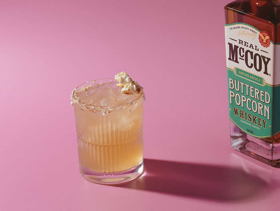 Whiskey bottle labeled "Real McCoy Buttered Popcorn Whiskey" beside a cocktail with a popcorn garnish, set on a pink background.