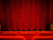 An empty theatre with rows of red seats facing a stage framed by red curtains