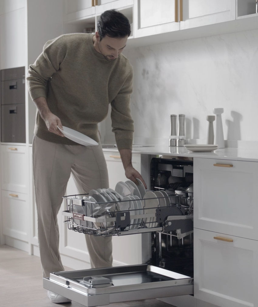 Man in a modern white kitchen pulls out a dishwasher rack full of plates while holding a dish.