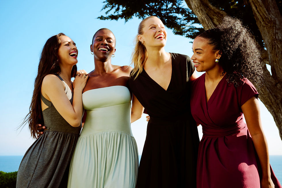 Four women in colorful dresses laugh together outdoors under a clear blue sky, with the ocean visible in the background. Joyful mood.