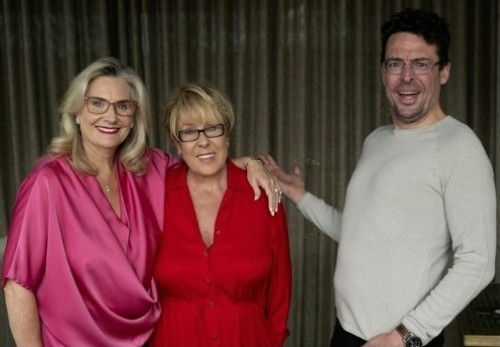 Three people smiling indoors; a woman in pink, a woman in red, and a man in white. Dark curtain backdrop. Warm, friendly atmosphere.
