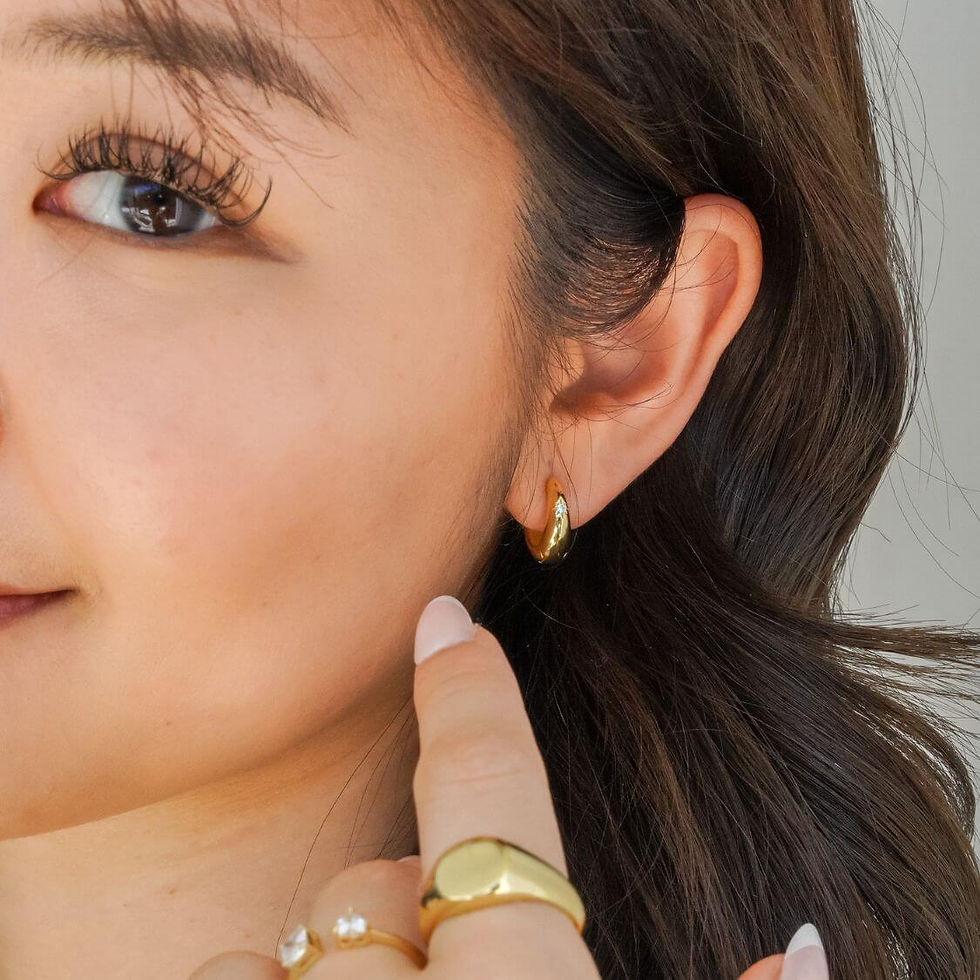 Close-up of a woman's face, highlighting a gold earring. She's pointing to it, with a gold ring visible on her finger, set against a neutral background.
