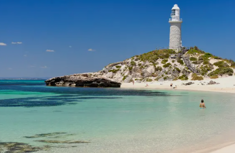 Beach scene with a white lighthouse on a hill. Clear turquoise water, people swimming and sunbathing, with a bright blue sky above.