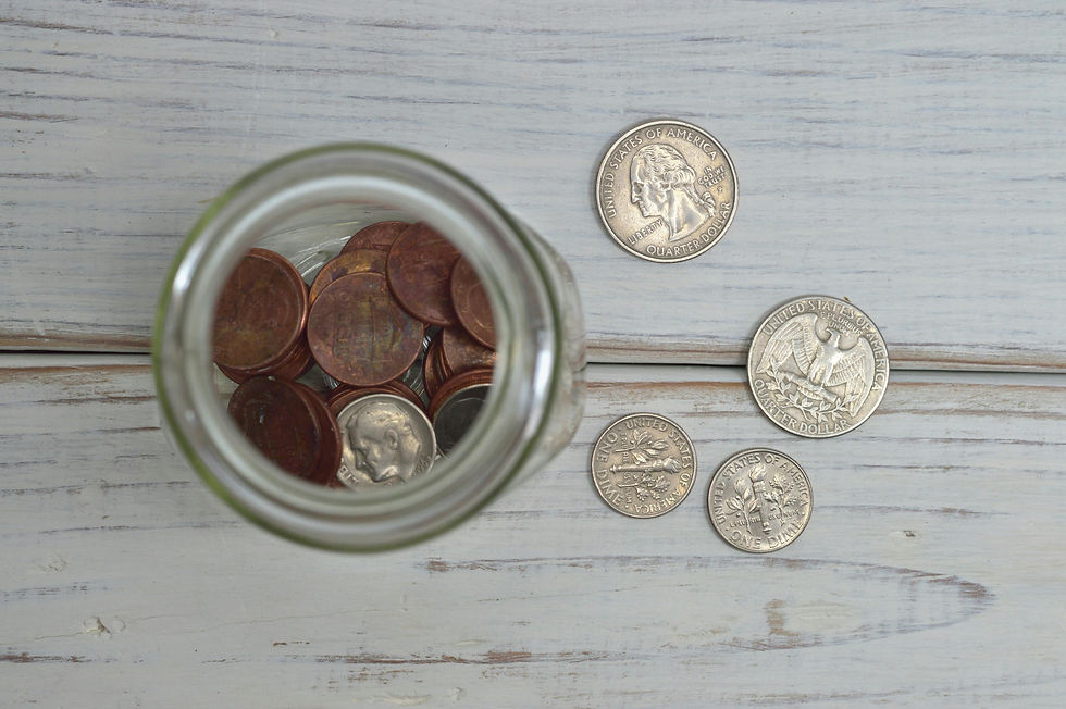 Glass jar filled with pennies and dimes on a wooden table, surrounded by spread-out quarters and dollar coins. Rustic setting.