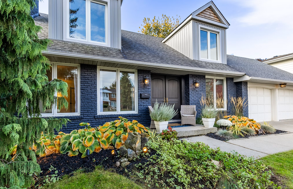 Blue brick suburban house with dormer windows, potted plants, and colorful front garden beside a driveway.