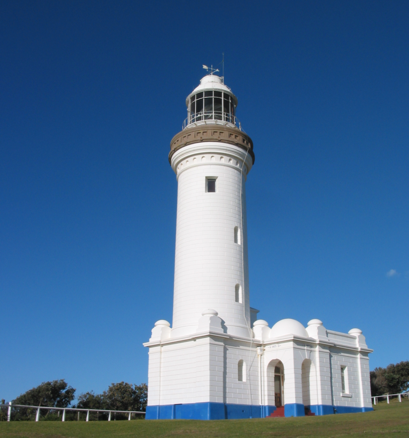 White lighthouse with a brown top stands against a bright blue sky. Green grass and trees surround the base, creating a serene atmosphere.