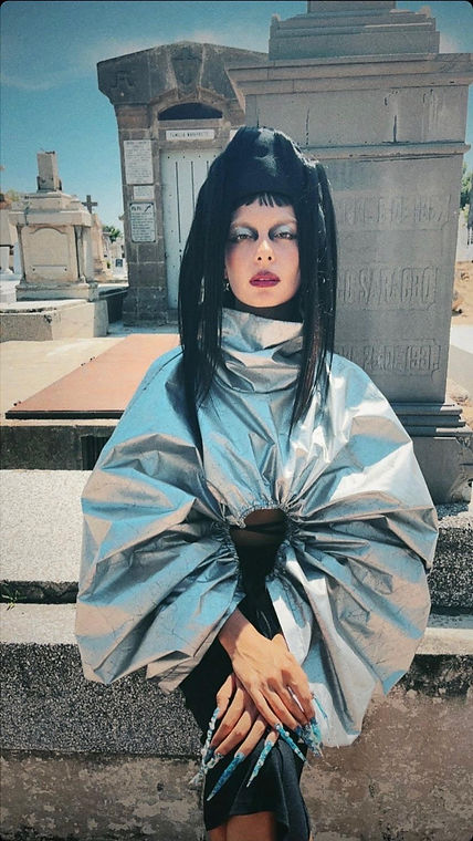 Una mujer vestida con un top metálico, uñas largas azules y sombrero negro con cabello sentada frente a una tumba.