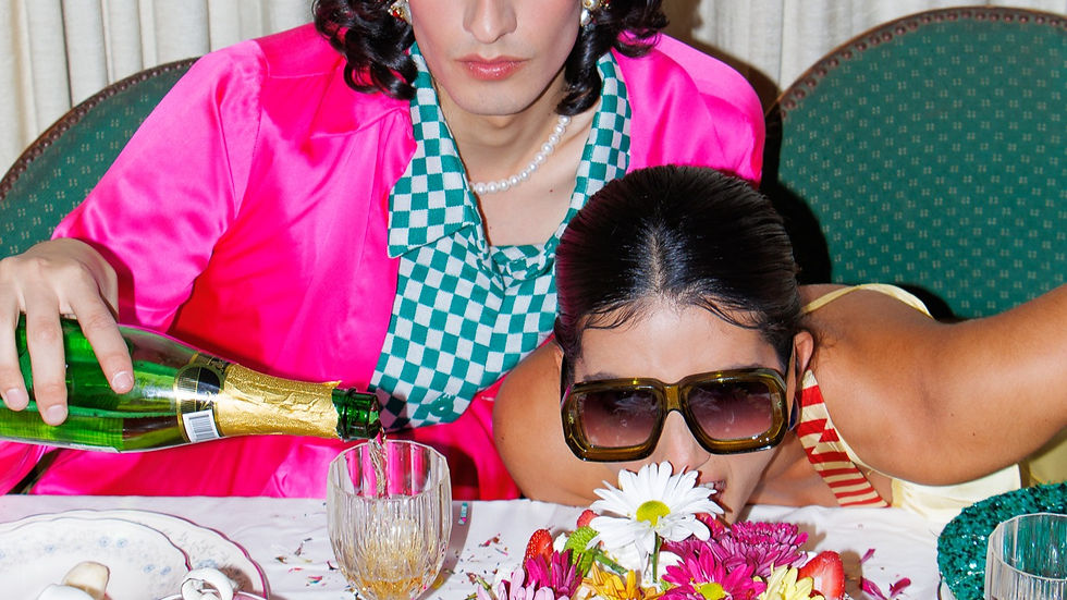 Fotografía editorial de un hombre con camisa rosa sirviendo champaña y una chica con lentes grandes mordiendo un pastel con flores