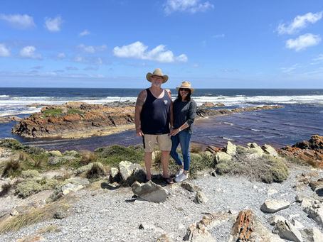 Jennifer and Bob standing together on rocky coastal terrain, ocean waves breaking in the background, under a partly cloudy sky.