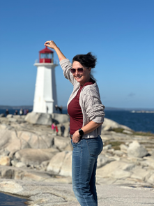 Smiling woman in sunglasses poses on rocky shore, arm raised, with a red-and-white lighthouse and blue sea behind her.