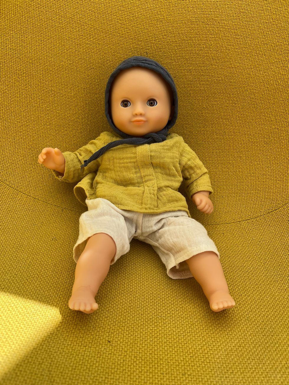 a cute doll from djeco in genderneutral coloured clothes sitting on a mustard yellow chair